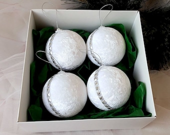 Puede incluir: Cuatro adornos navideños de terciopelo blanco con ribetes plateados en una caja de regalo blanca forrada con papel de seda verde.
