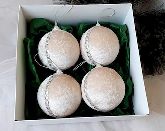 Puede incluir: Cuatro adornos navideños de terciopelo color crema con ribetes plateados en una caja blanca con papel de seda verde.
