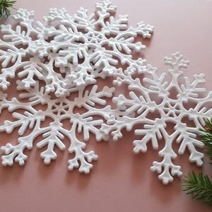 May include: White snowflake ornaments with intricate designs are arranged on a pink surface, with green pine branches in the corners. These decorative snowflakes are perfect for winter holiday decor.