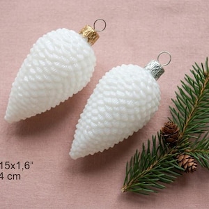 May include: Two white, textured, cone-shaped Christmas ornaments with gold and silver caps. A sprig of green pine needles and small pine cones are next to the ornaments. The ornaments are 3.15 x 1.6 inches (8x4 cm).