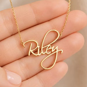 Pode incluir: Um colar de tom dourado com uma corrente fina e um pingente que forma o nome Riley escrito em letra cursiva fluida e contínua. O metal tem um acabamento polido e as letras estão conectadas em uma única linha elegante.