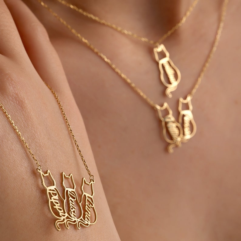 Pu&ograve; includere: Collana in oro con tre silhouette di gatti, ciascuna con un nome inciso: "Lucy", "Daisy" e "Harper".