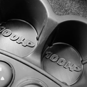 Könnte beinhalten: Schwarz-Weiß-Nahaufnahme eines Getränkehalters im Auto. Zwei runde Einsätze mit dem erhabenen Text "100HP" sind zu sehen. Der Getränkehalter ist aus dunklem Kunststoff gefertigt, mit einer stilisierten Schriftart.