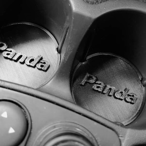 Könnte beinhalten: Schwarz-Weiß-Bild von zwei Getränkehaltern im Auto mit individuellen Einsätzen. Jeder Einsatz trägt das Wort "Panda" in erhabenen Buchstaben. Die Getränkehalter sind Teil der Konsole eines Autos, darunter ist ein Bedienfeld zu sehen.
