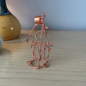 May include: A copper wire sculpture shaped like a cello with a bow positioned across the strings. The metallic wire is bent into the outline of the musical instrument, featuring a scroll at the top, a body with f-holes, and a bridge.