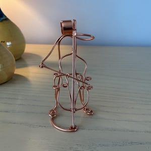 May include: A copper wire sculpture depicting a musician playing a double bass. The figure is constructed from thin, bent copper wire, forming the outline of a person seated while holding and playing a stringed instrument with a bow.