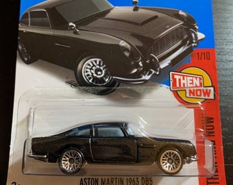 May include: A black 1963 Aston Martin DB5 Hot Wheels die-cast car in a plastic package. The car is a 1/10 scale model and is part of the "Then & Now" series. The package also includes a QR code for the "Showdown" app.