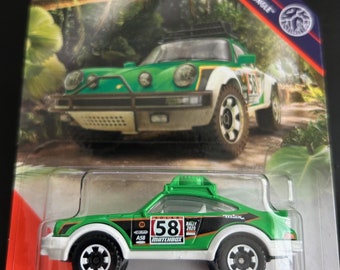 Könnte beinhalten: Ein Matchbox-Spielzeugauto, ein grüner und weißer '85 Porsche 911 Rally. Das Spielzeugauto hat schwarze Räder und die Nummer 58 auf der Seite. Die Verpackung zeigt eine Dschungelszene und das Matchbox-Logo.