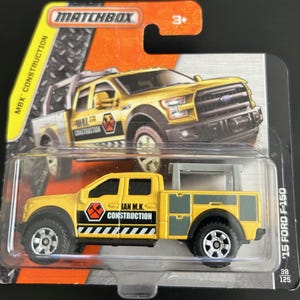 Könnte beinhalten: Gelber Matchbox '15 Ford F-150 Spielzeug-LKW mit einem schwarz-gelben Bauschild mit der Aufschrift "Ian M.K. Construction". Der LKW hat eine graue Werkzeugkiste in der Ladefläche und ein graues Leitergestell auf der Kabine.