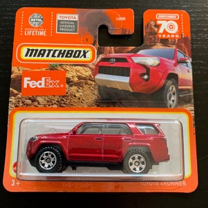Peut inclure: Voiture miniature rouge Toyota 4Runner Matchbox avec la marque FedEx sur le côté. La voiture est sur un fond blanc avec une bordure rouge et orange. L'emballage comprend le logo Matchbox et les mots "70 ans".