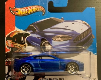 May include: A blue die-cast toy car model of an Aston Martin DBS, part of the Hot Wheels collection. The car is on a white background with a red and black graphic in the background. The car is numbered 153/250.