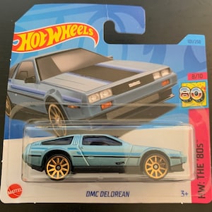 May include: A Hot Wheels die-cast model of a DMC DeLorean car. The car is light blue with gold wheels and black accents. The packaging features the Hot Wheels logo and the text "HW: THE '80S".