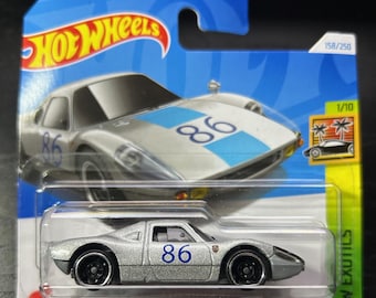 Könnte beinhalten: Ein silbernes Hot Wheels-Spielzeugauto aus Metallguss, ein Porsche 904 Carrera GTS, mit einem blauen Rennstreifen und der Nummer 86 an der Seite. Das Auto befindet sich auf einem blauen und gelben Hintergrund mit dem Hot Wheels-Logo und den Worten "HW Exotics".
