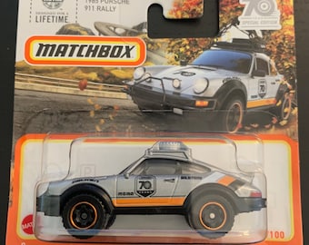 Könnte beinhalten: Ein silbernes Matchbox 1985 Porsche 911 Rally Spielzeugauto mit schwarzen Akzenten, orangefarbenen Rädern und einem 70-jährigen Jubiläumslogo. Das Auto befindet sich auf einem orangefarbenen und weißen Hintergrund mit dem Matchbox-Logo.