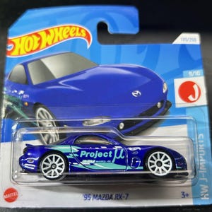 May include: A blue 1995 Mazda RX-7 die-cast toy car with white wheels and "Project μ" printed on the side. The car is part of the Hot Wheels J-Imports series.