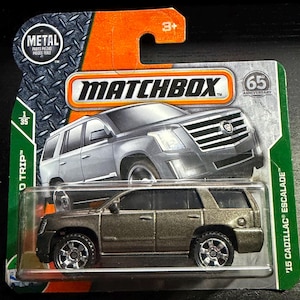 May include: A Matchbox die-cast model of a '15 Cadillac Escalade. The toy car is metallic brown with black wheels and sits on a green and orange cardboard backing. The packaging reads "Matchbox" and "65 Anniversary".