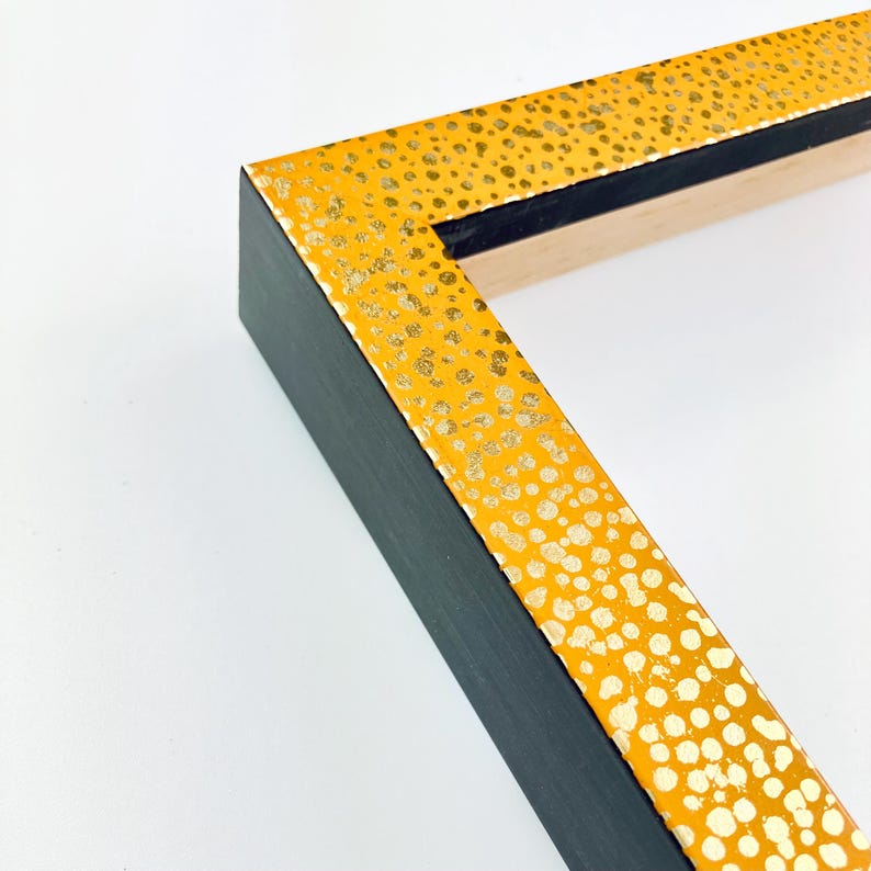 May include: A close-up of a picture frame with a black wood frame and a yellow border with gold flecks. The frame is angled to show the corner.