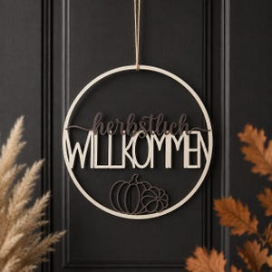 Könnte beinhalten: Ein kreisförmiger Wandbehang aus Holz mit dem Schriftzug "herbstlich" in einer dunklen Schreibschrift über dem Wort "WILLKOMMEN" in einer hellen Blockschrift. Unter dem Text befinden sich zwei dunkle Kürbissilhouetten im Holzrahmen.