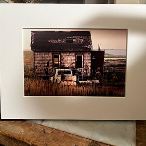 May include: A framed print of a weathered house and an old truck in a field. The house has a damaged roof and wooden siding. The truck is rusty and sits in tall grass. The sky is a pale pink.