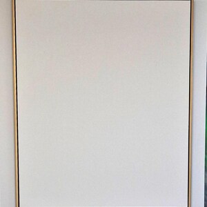 May include: A square, blank canvas with a white surface, framed by a thin black border and a gold-coloured outer frame. The artwork is mounted on a white wall.