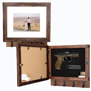 May include: A dark wood wall-mounted display case featuring a hinged picture frame front. The interior contains a tan-colored handgun and a black magazine set into black foam. The exterior frame includes four black metal hooks for hanging items.