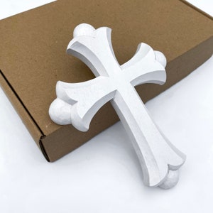 May include: White wooden cross with a detailed, carved design. The cross has a rounded, ornate shape at the top and bottom.
