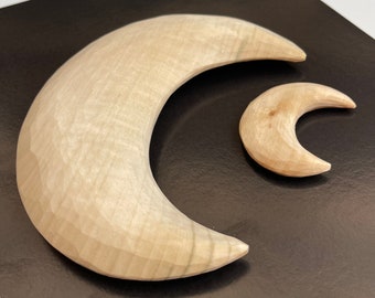 Könnte beinhalten: Zwei hölzerne Halbmondformen, eine große und eine kleine, werden vor einem dunklen Hintergrund gezeigt. Die Monde sind aus hellem Holz geschnitzt und zeigen die natürliche Maserung und Textur. Der größere Mond ist etwa 20 cm lang, während der kleinere etwa 8 cm misst.