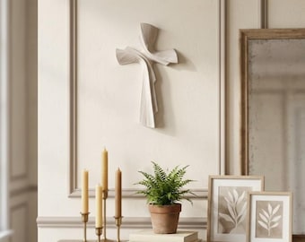 Peut inclure: Une sculpture murale en forme de croix avec une finition blanc cassé lisse, présentant des lignes fluides, superposées et courbes qui créent un mouvement. La croix possède une surface texturée avec de fines stries et un design épuré.