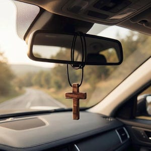 May include: A wooden cross car charm hanging from the rearview mirror. The cross is a medium brown color and is suspended by a black cord. The car's dashboard is black, and the background shows a road and trees.