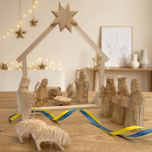 Op de afbeelding: Een houten kerststalset met uitgesneden figuren van mensen en dieren, opgesteld onder een eenvoudig frame met een ster aan de bovenkant. De set bevat een kribbe, schapen en figuren met geschenken, geplaatst op een blauw en geel lint.