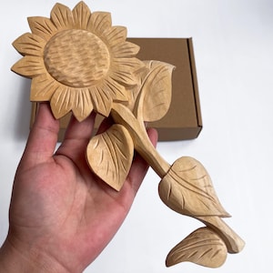 Puede incluir: Un girasol de madera tallado a mano con pétalos, hojas y tallo detallados. El girasol es de color madera claro y natural. La talla se sostiene en una mano, con una caja de cartón marrón en el fondo.