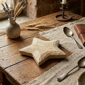 Op de afbeelding: Een houten stervormig decoratief object met gesneden details. De ster is licht van kleur en staat op een houten tafel, naast een kleine vaas met gedroogde bloemen, lepels en een leren boek.