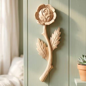 Puede incluir: Decoración de pared de madera tallada que representa una rosa en plena floración con pétalos y hojas detalladas. La pieza está montada sobre un panel de madera verde claro. La madera tiene un aspecto natural sin terminar.