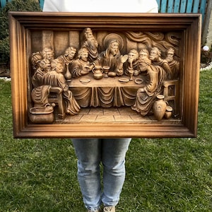 May include: A detailed, brown wooden relief carving depicting the Last Supper. The artwork features figures seated around a table, with intricate details in their clothing and expressions. The carving is framed in a dark brown, rectangular wooden frame.
