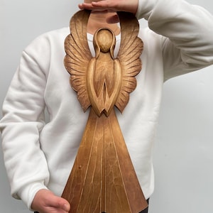 May include: A wooden angel sculpture with outstretched wings and a flowing gown. The angel is carved from light brown wood, with detailed feather and garment textures. The sculpture is approximately 61 cm tall.