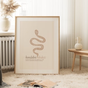 Könnte beinhalten: Ein gerahmter Druck mit beigefarbenem Hintergrund, der eine stilisierte Illustration einer Schlange mit dem Text "kundalini shakti" und einer Definition darunter zeigt. Der Druck ist in einem hellen Holzrahmen gefasst und hängt an einer Wand.