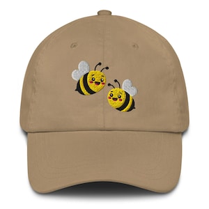 Könnte beinhalten: Eine khakifarbene Baseballkappe mit einer Stickerei von zwei kleinen, lächelnden Bienen. Jede Biene hat gelb-schwarze Streifen, weiße Flügel und rosa Wangen. Die Kappe verfügt über einen gebogenen Schirm und eine strukturierte Krone mit Knopf.