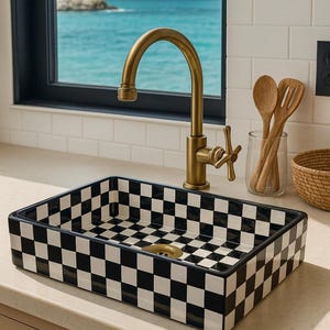 Puede incluir: Un fregadero rectangular a cuadros blancos y negros con un grifo de latón. El fregadero está situado frente a una ventana con vistas al océano. Utensilios de madera en un tarro de cristal.