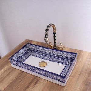 Könnte beinhalten: Ein rechteckiges Badezimmerwaschbecken aus weißer Keramik mit einem blauen und weißen geometrischen Muster am Rand. Das Waschbecken steht auf einer Holzarbeitsplatte mit einem goldenen Wasserhahn mit einem schwarz-weißen Muster am Griff.