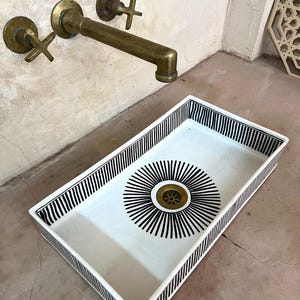 Op de afbeelding: Een rechthoekige witte keramische gootsteen met zwart-wit gestreepte rand. De gootsteen heeft een zwart-wit zonnestralenontwerp rond de afvoer.