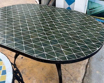 Puede incluir: Una mesa de café ovalada con un mosaico de azulejos verdes y blancos y un marco y patas de metal negro. La mesa está sobre una superficie de hormigón.