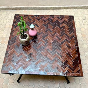 Può includere: Tavolino quadrato con piano a mosaico a motivi marroni, disposti a spina di pesce. Il tavolo ha gambe in metallo nero. Una piccola pianta in vaso e un vaso rosa con la parte superiore blu sono sul tavolo.