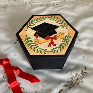 May include: A black hexagonal box with a graduation cap and diploma design. The box is decorated with green leaves and a red ribbon. The box is sitting on a white fabric surface with gold embroidery.