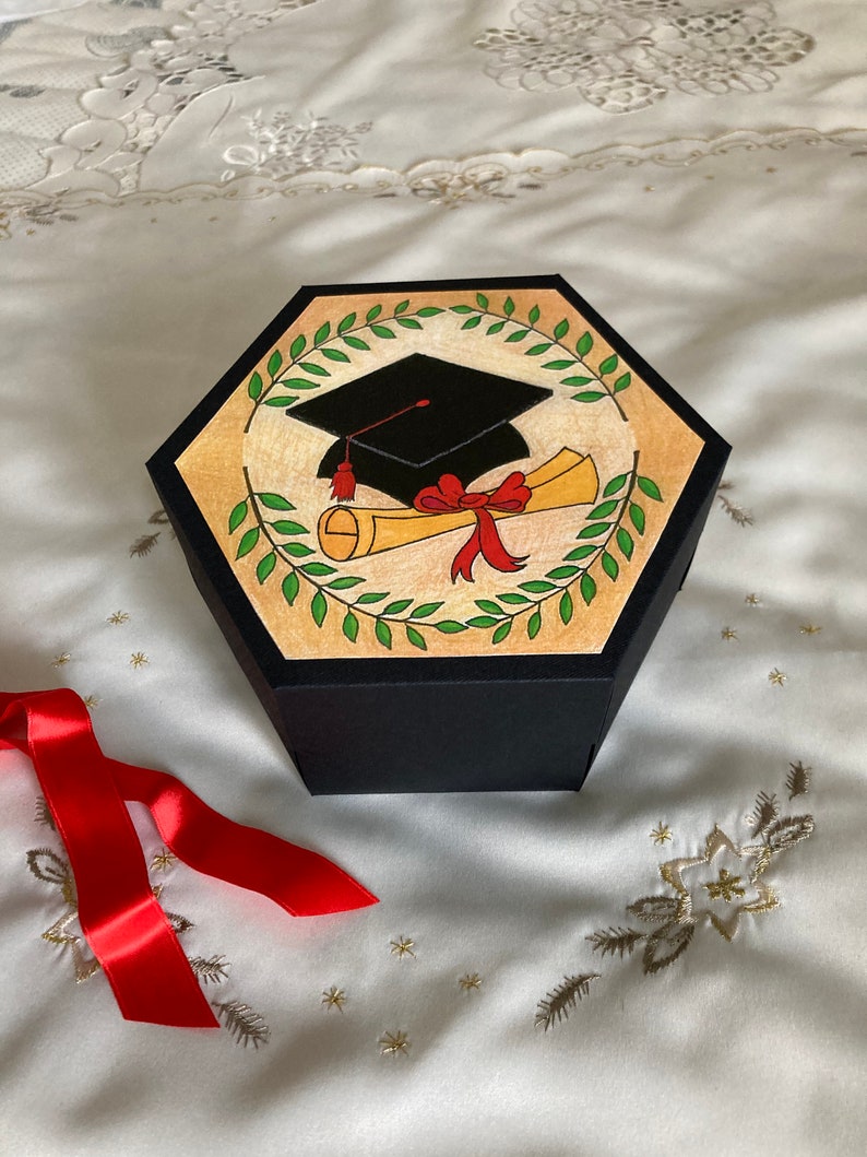 May include: A black hexagonal box with a graduation cap and diploma design. The box is decorated with green leaves and a red ribbon. The box is sitting on a white fabric surface with gold embroidery.