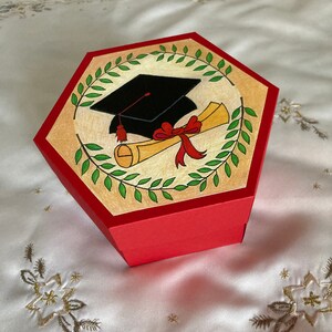 May include: Red hexagonal box with a graduation cap and diploma illustration. The illustration is surrounded by a green wreath. The box is closed.
