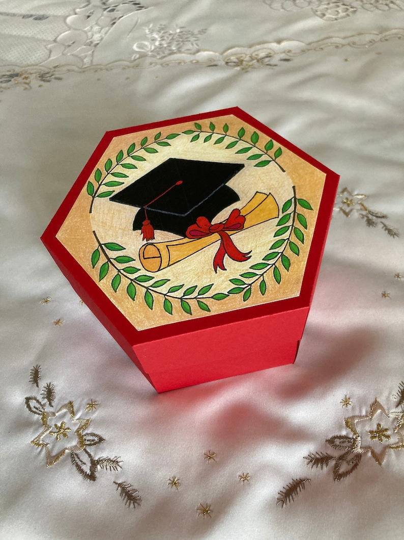 May include: Red hexagonal box with a graduation cap and diploma illustration. The illustration is surrounded by a green wreath. The box is closed.