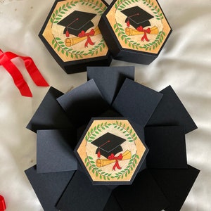 May include: Two black hexagonal boxes with a graduation cap and diploma design. The boxes are open to reveal a larger black box with multiple flaps. The flaps are folded to create a multi-layered box.