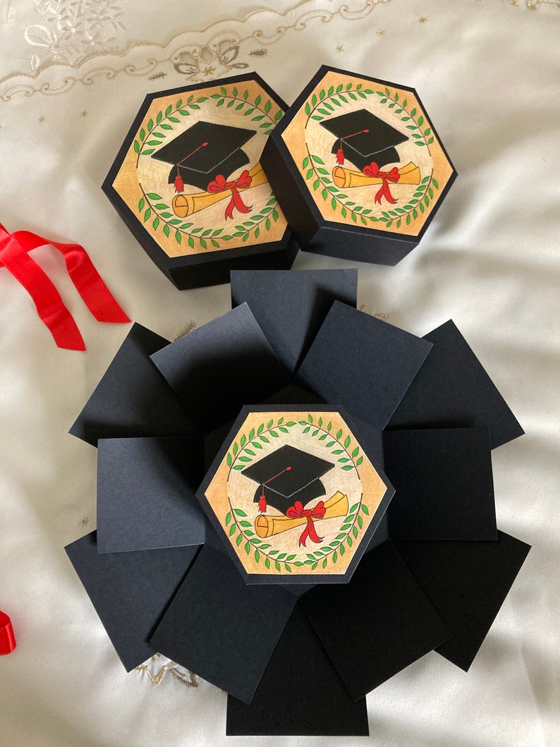 May include: Two black hexagonal boxes with a graduation cap and diploma design. The boxes are open to reveal a larger black box with multiple flaps. The flaps are folded to create a multi-layered box.