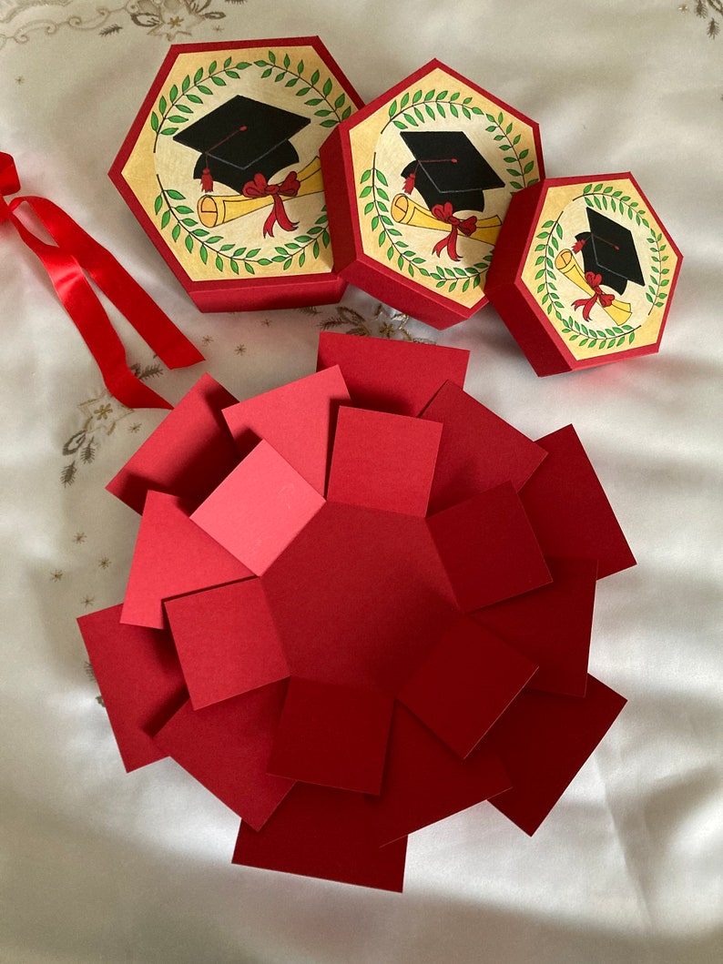 May include: Three red and white graduation themed gift boxes with a black graduation cap and diploma design. The boxes are open and folded flat, revealing a red paper hexagon.