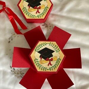 May include: A red hexagonal box with a graduation cap and diploma design. The box is open and the sides are folded out to reveal the interior.